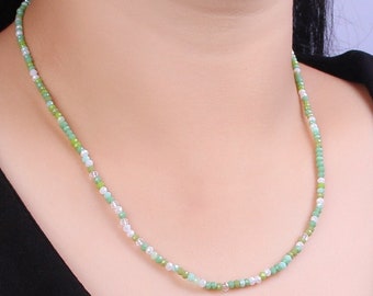 May include: A delicate necklace composed of small, round beads in shades of green, white, and pale pink. The necklace is worn against a black top, highlighting the colors of the beads. The beads appear to be strung on a thin, flexible cord.
