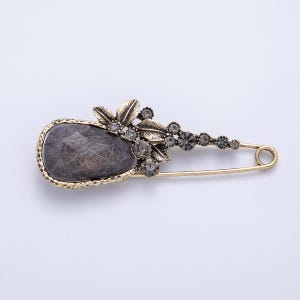 May include: A gold-toned safety pin brooch with a large, gray, teardrop-shaped stone at the center. The stone is surrounded by a gold-colored frame and adorned with small, gray crystals and gold-colored leaves. The pin is a decorative accessory.