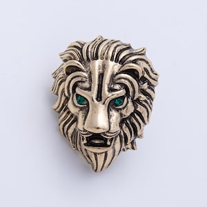 May include: A gold-colored lion head brooch with green gemstone eyes. The brooch features detailed mane and facial features, set against a white background. This decorative pin is suitable for jackets, bags, or hats.