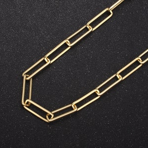Puede incluir: Un collar de cadena dorado con eslabones rectangulares. La cadena se muestra sobre un fondo oscuro y texturizado, resaltando su apariencia brillante y metálica. El collar es una pieza de joyería sencilla y elegante.