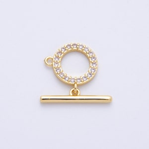 Puede incluir: Un cierre de palanca dorado con un anillo redondo con cristales y una barra recta. El anillo tiene un pequeño lazo. El cierre está diseñado para la fabricación de joyas y la artesanía.