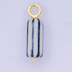 以下が含まれることがあります： 金色のペンダントで、円筒形のストライプビーズが付いています。ビーズは白と青の縦縞模様で、上部に金色のループ、端に金色のキャップが付いています。ペンダントはジュエリー作りのために設計されています。