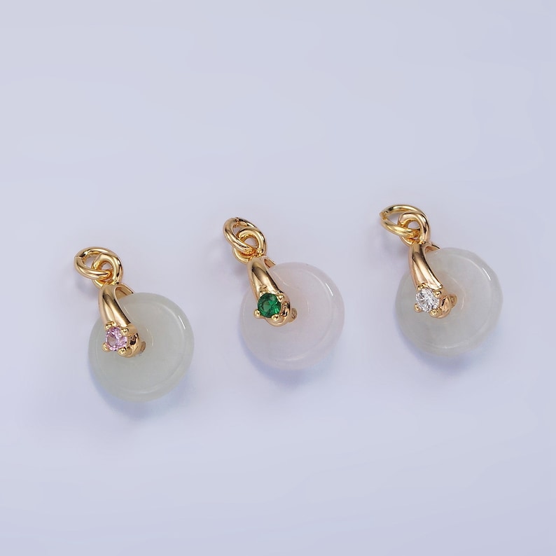 Pu&ograve; includere: Tre ciondoli dorati con un disco di giada bianca e una piccola pietra preziosa al centro. I ciondoli sono di forma circolare con un anello dorato in alto. Le pietre preziose sono rosa, verde e trasparente.