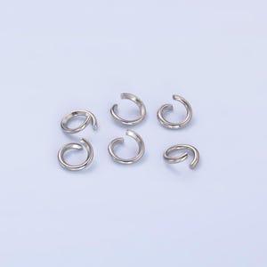 May include: Six silver jump rings, also known as split rings, are arranged on a light blue background. These rings are commonly used in jewelry making and crafting.