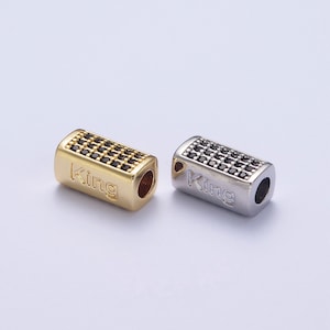 May include: Two rectangular metal beads with black cubic zirconia stones and the word "King" engraved on the side. One bead is gold and the other is silver.