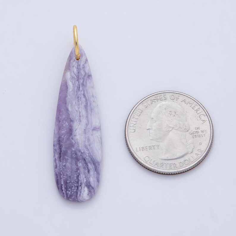 May include: A teardrop-shaped pendant with a gold-colored bail. The pendant is a marbled lavender and white stone. A U.S. quarter is next to the pendant for size comparison. The pendant is likely for jewelry.