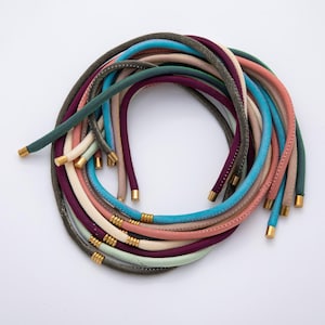 May include: A collection of colorful necklaces in various shades, including teal, pink, purple, and gray. Each necklace features a cord design with gold-colored metal ends. The necklaces are arranged in a circular pattern against a white background.