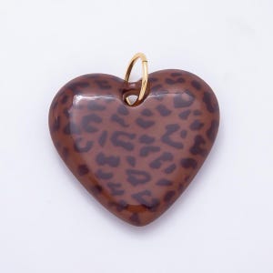 Può includere: Un ciondolo a forma di cuore con una superficie marrone lucida e un motivo leopardato marrone scuro. Il ciondolo include un piccolo anello di giunzione circolare in metallo dorato fissato al centro superiore per appenderlo o collegarlo.
