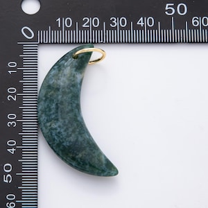 Op de afbeelding: Een maanvormige hanger van groen en wit gevlekte steen. Het heeft een goudkleurige ring aan de bovenkant om op te hangen. De hanger is ongeveer 5 cm lang.