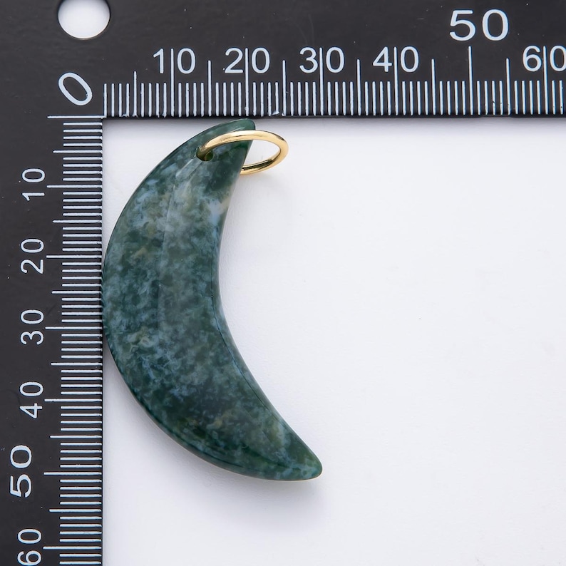 Op de afbeelding: Een maanvormige hanger van groen en wit gevlekte steen. Het heeft een goudkleurige ring aan de bovenkant om op te hangen. De hanger is ongeveer 5 cm lang.