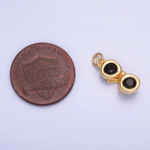 May include: A gold-colored charm shaped like binoculars, featuring two black gemstones. The charm is next to a United States one-cent coin for size comparison. The charm has a small loop at the top for attaching to a necklace or bracelet.