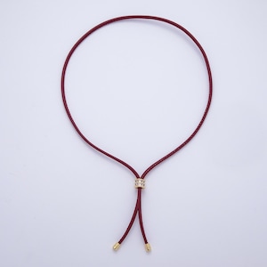 May include: A burgundy leather necklace with a gold-colored accent and gold-colored tips. The necklace has a circular design with a sliding gold-colored accent in the center. The necklace is displayed on a white background.