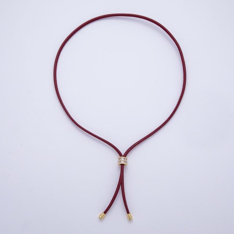 May include: A burgundy leather necklace with a gold-colored accent and gold-colored tips. The necklace has a circular design with a sliding gold-colored accent in the center. The necklace is displayed on a white background.