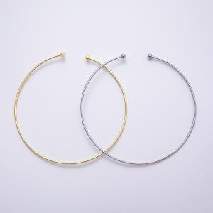 May include: Two minimalist choker necklaces. One is gold-toned, the other is silver-toned. Each necklace features a thin, curved band that forms a circular shape, with a small ball at each end. The necklaces are displayed on a white background.