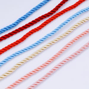 May include: Close-up of six colorful twisted ropes in shades of blue, red, light blue, cream, and pink. The ropes are diagonally arranged, showcasing their textured, spiraled design. The background is a plain white.
