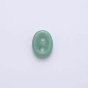 May include: A light green, oval-shaped button with two holes. The button is made of a smooth, possibly stone-like material. The button is centered on a white background.