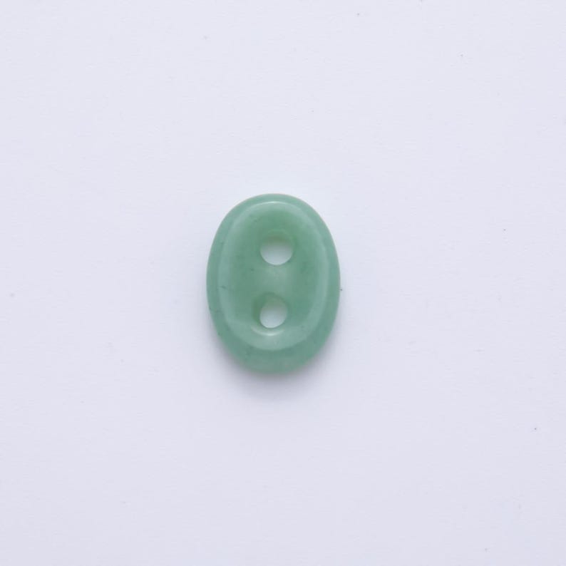 May include: A light green, oval-shaped button with two holes. The button is made of a smooth, possibly stone-like material. The button is centered on a white background.