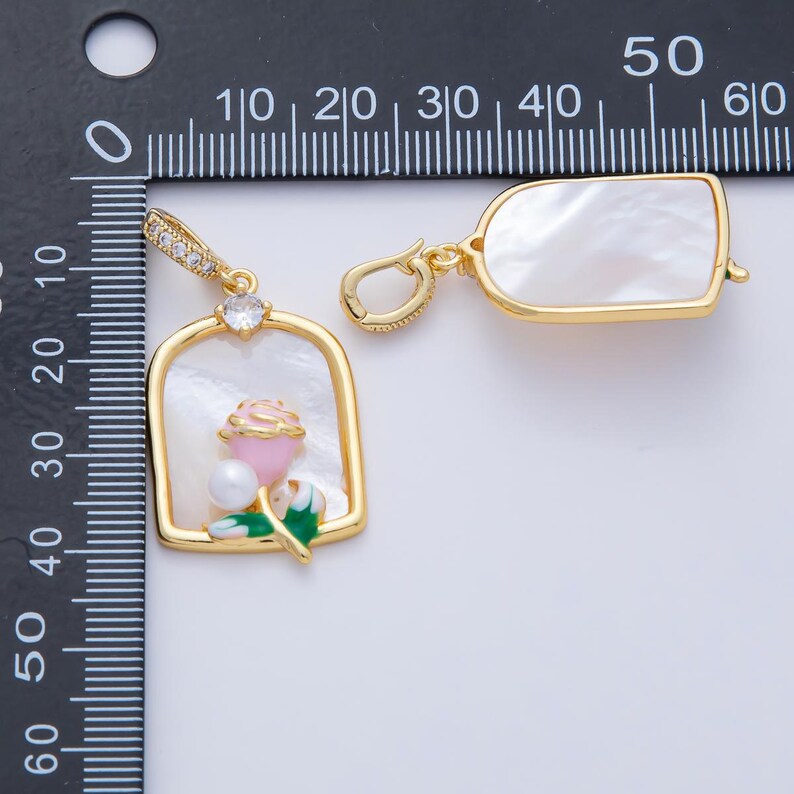 May include: Two gold-toned pendants with white mother-of-pearl inserts. One pendant features a pink rose with a pearl and green leaves. The other pendant has a rectangular mother-of-pearl insert. Both pendants have a decorative bail.