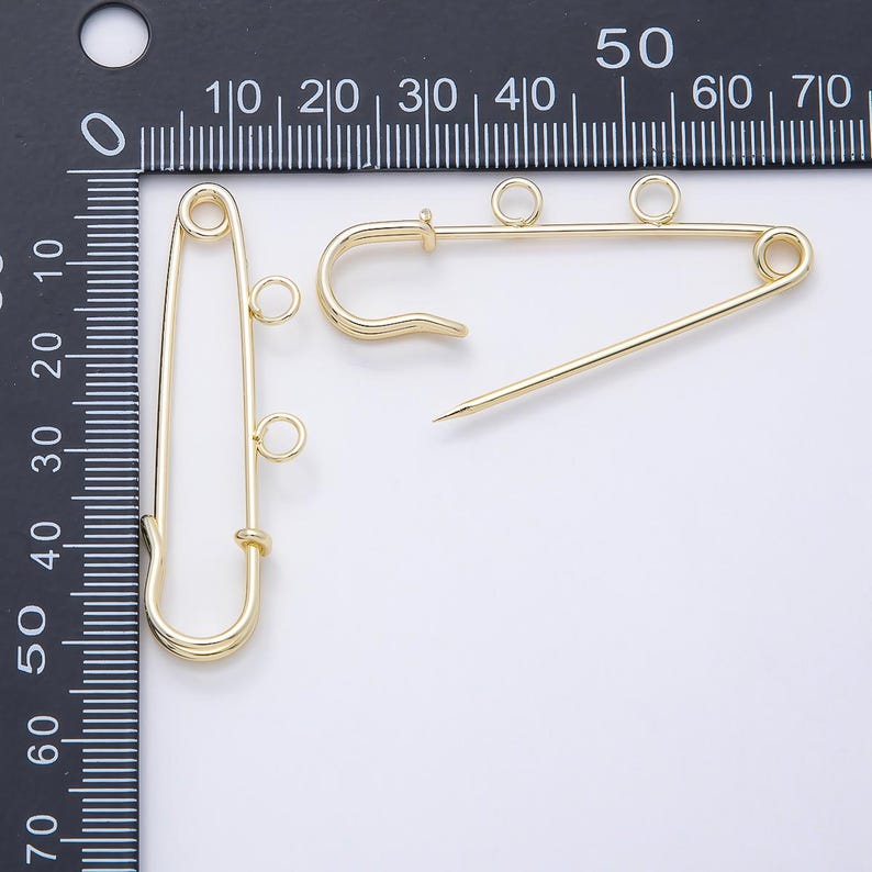 May include: Two gold-toned safety pins, each featuring two small circular loops attached to the side. The pins are positioned against a black ruler with white markings, showing a length of approximately 2.25 inches for the vertical pin.