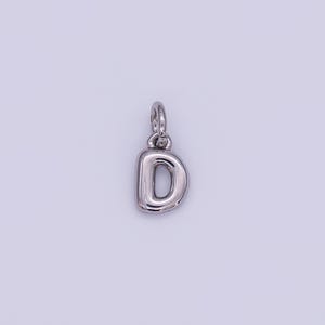 May include: A silver-colored pendant in the shape of the letter "D". The pendant has a rounded, bubble-like design and includes a small loop at the top for attaching to a chain or bracelet. The background is a light, neutral color.