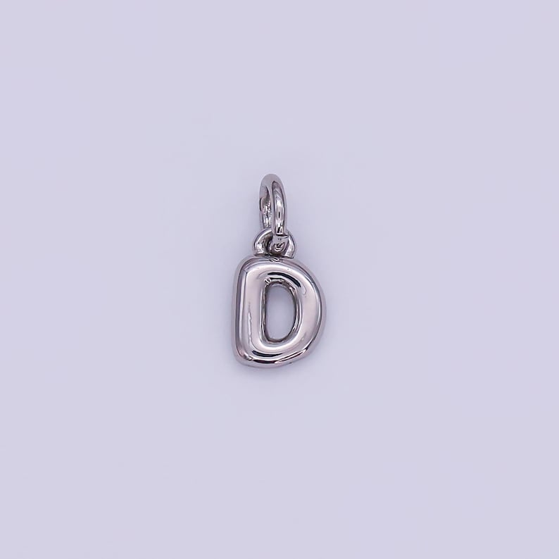 May include: A silver-colored pendant in the shape of the letter "D". The pendant has a rounded, bubble-like design and includes a small loop at the top for attaching to a chain or bracelet. The background is a light, neutral color.