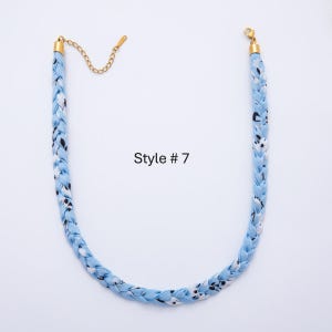 May include: A braided necklace in light blue fabric with a pattern of white and black shapes. The necklace has gold-colored metal clasps and chain. The text "Style # 7" is printed on the image.