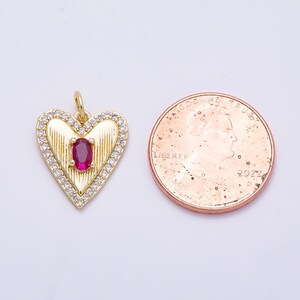 May include: A gold-toned heart-shaped pendant featuring a central oval-cut red stone, vertical ribbed detailing, and a border of small clear stones, positioned next to a 2022 United States penny for scale to show its size.