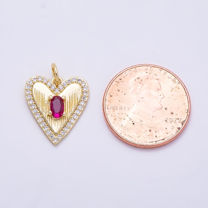 May include: A gold-toned heart-shaped pendant featuring a central oval-cut red stone, vertical ribbed detailing, and a border of small clear stones, positioned next to a 2022 United States penny for scale to show its size.