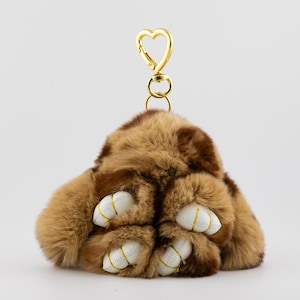 May include: A plush, brown and tan paw-shaped keychain with white paw pads and gold stitching. The keychain has a gold heart-shaped clasp for easy attachment. The faux fur is soft and fluffy, creating a playful accessory.