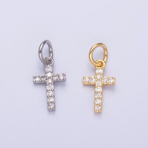 May include: Two cross pendants, one silver-toned and one gold-toned, each encrusted with clear, sparkling gemstones. The crosses have a simple, elegant design, perfect for religious jewelry or crafting. The pendants are shown against a light purple background.