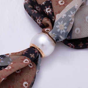 May include: A scarf ring featuring a large, smooth, white faux pearl bead with a gold-toned metal base, holding a lightweight fabric scarf patterned with a repeating floral design in shades of brown, black, and muted blue.