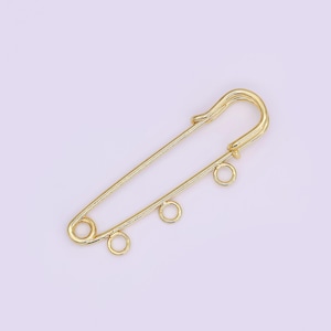 May include: A gold-colored safety pin with three small, round loops attached. The pin is a simple, elegant design, perfect for jewelry making or crafting. The pin is shown against a light purple background.