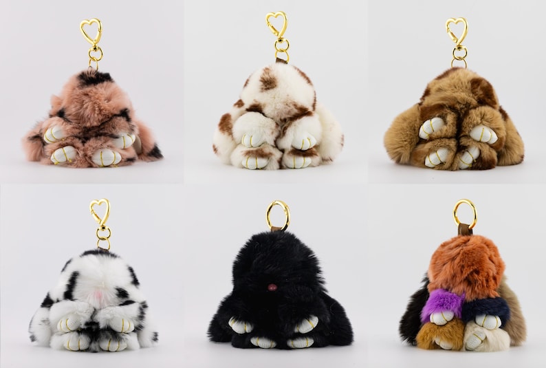 May include: Six fluffy bunny-shaped keychains in various colors. The keychains have gold-colored heart-shaped clasps. Colors include pink and brown, brown and white, tan and brown, black and white, solid black, and a mix of colors.