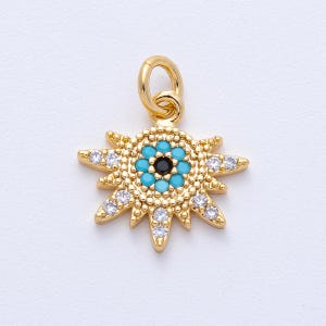 May include: Gold-colored charm with a sunburst design. The center features a turquoise and black eye-like pattern, surrounded by clear, sparkling accents. The charm has a loop at the top for attachment.