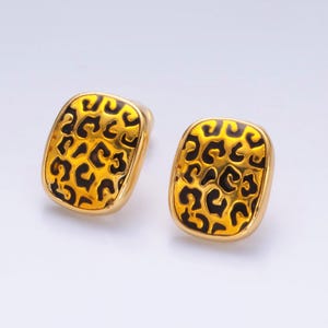 Puede incluir: Un par de pendientes cuadrados dorados. Cada pendiente presenta un diseño de estampado de leopardo negro sobre un fondo dorado. Los pendientes tienen forma cuadrada redondeada y cierre de poste.