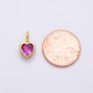 May include: A gold-toned heart-shaped pendant featuring a faceted pink gemstone center, positioned next to a United States one-cent copper coin for scale, showing the relative size of the jewelry piece against the 0.75 inch diameter coin.