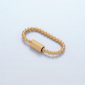 Może przedstawiać: Złota bransoletka z koralików z cylindrycznym wisiorkiem. Wisiorek ma fakturowaną powierzchnię.