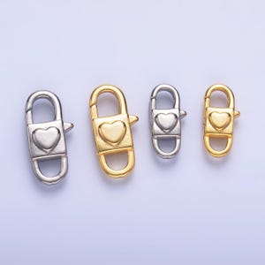 May include: Four metal lobster clasps in silver and gold tones. Each clasp features a heart-shaped design in the center. The clasps are in varying sizes, with a light purple background.