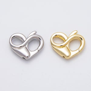 May include: Two heart-shaped lobster clasps, one silver-colored and one gold-colored, are displayed against a white background. The clasps have a smooth, polished finish and a curved design, ideal for jewelry making or crafting.