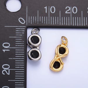 May include: Two pendants, one silver-toned and one gold-toned, each featuring two black gemstones. The silver pendant is a simple design, while the gold pendant has an infinity symbol shape. The pendants are next to a ruler.