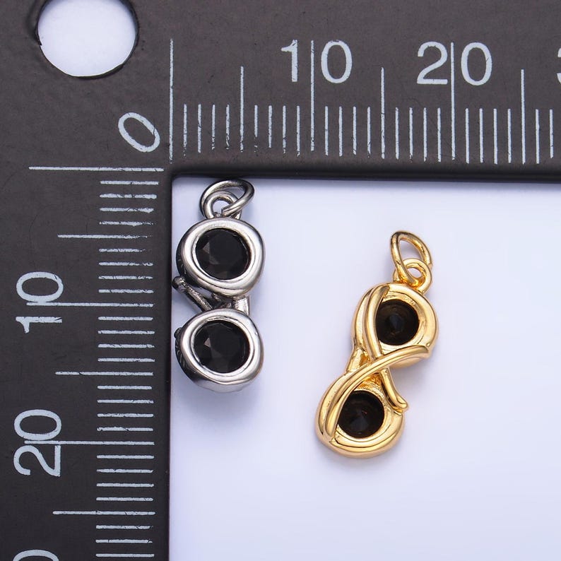 May include: Two pendants, one silver-toned and one gold-toned, each featuring two black gemstones. The silver pendant is a simple design, while the gold pendant has an infinity symbol shape. The pendants are next to a ruler.