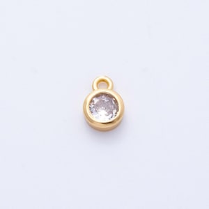 May include: A small, gold-colored charm with a round, clear gemstone set in the center. The charm has a loop at the top for attaching to a necklace or bracelet. The gemstone sparkles against the gold setting.