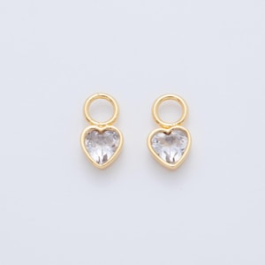 May include: Two gold-colored heart-shaped charms with clear, faceted centers. Each charm has a small loop at the top for attachment. The charms are set against a plain white background.