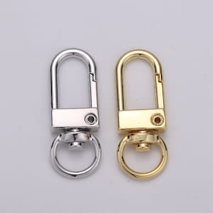May include: Two metal swivel clasps, one silver and one gold, are displayed against a light background. Each clasp has a rectangular body with a circular ring at the bottom and a curved top that opens and closes.