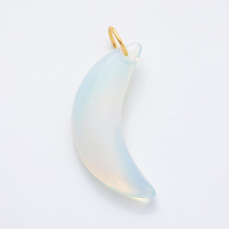 Op de afbeelding: Een maanvormige hanger met een goudkleurige lus. De hanger heeft een bleke, doorschijnende blauw-witte kleur. De maanvorm is glad en gebogen, met een subtiele kleurovergang.