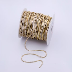 Puede incluir: Un carrete de cadena de strass dorado. La cadena está hecha de pequeños strass cuadrados que están engastados en un marco de metal. La cadena está enrollada alrededor de un carrete blanco.
