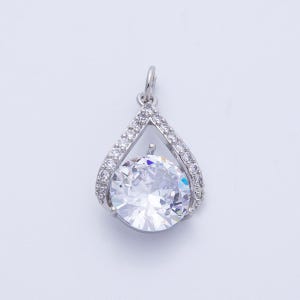 May include: A silver pendant with a large, clear, round gemstone at the center. The gemstone is surrounded by a teardrop-shaped frame encrusted with smaller, clear stones. The pendant has a small loop at the top for a chain.