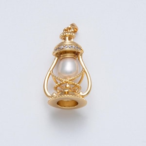 May include: A gold-colored lantern pendant with a pearl center and small, clear gemstones around the top. The pendant has intricate detailing and a bail for attaching to a chain. The lantern design is reminiscent of vintage or antique jewelry.
