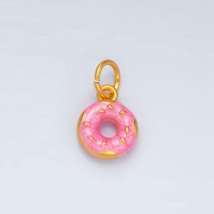 Könnte beinhalten: Ein goldfarbener Donut-Anhänger mit rosa Emaille und weißen Streuseln. Der Anhänger hat eine Öse oben zum Anbringen an einer Halskette oder einem Armband.