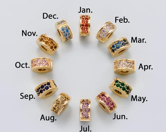 May include: A collection of gold-toned, cylindrical beads, each adorned with a band of colorful gemstones. The beads are arranged in a circle, with months of the year labeled around them, such as "Jan.", "Feb.", and "Mar."